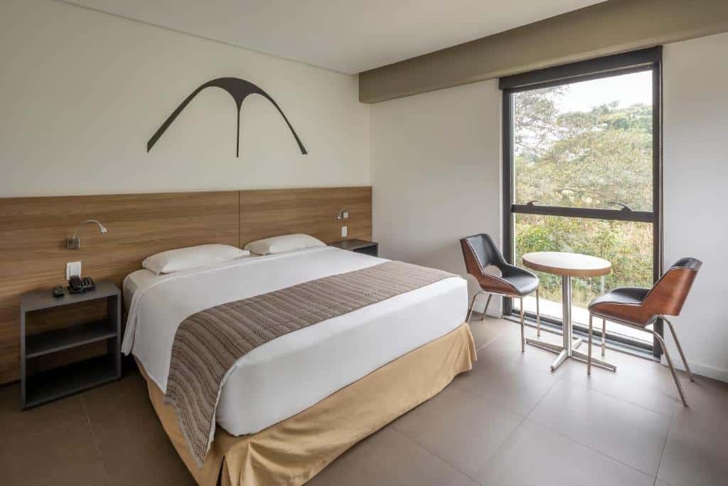 Quarto do Linx Galeão by Wish com uma cama de casal, duas cadeiras, uma mesa redonda e uma grande janela com árvores do lado de fora. Essa é a uma das opções de onde ficar no Rio de Janeiro.