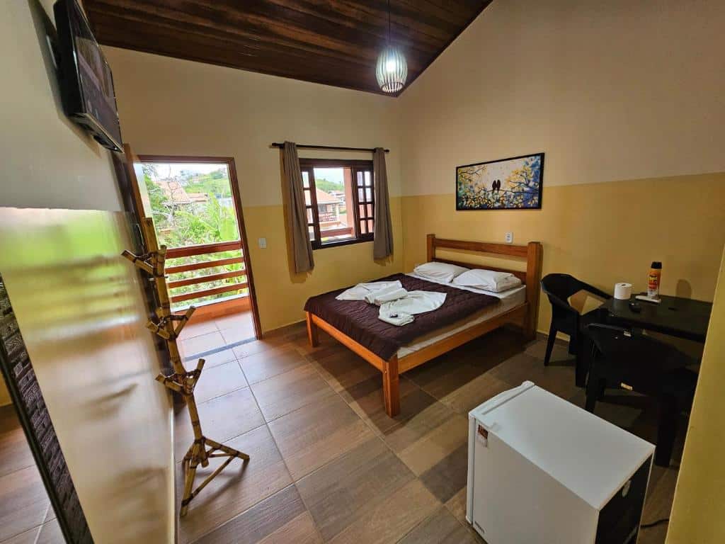 Um quarto simples com uma cama de casal, frigobar pequeno, mesa com cadeiras, cabide de parede, TV e porta da varanda.