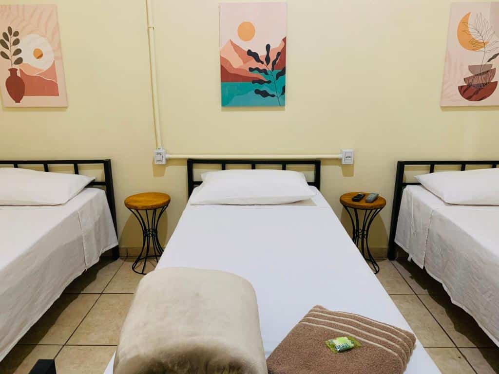 Pousada Quinzitão, com três camas de solteiro com lençóis brancos em um quarto, cada uma com uma mesa lateral; obras de arte penduradas na parede de cor creme.