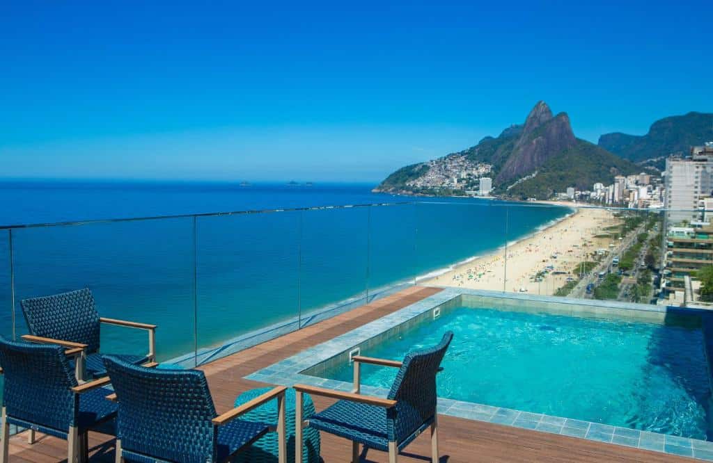 A piscina na cobertura do Sol Ipanema Hotelcom cadeiras tem vista para uma praia de areia e para o oceano, com montanhas e prédios da cidade ao fundo.