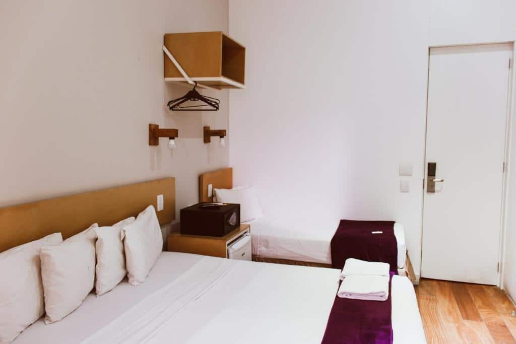 Quarto do Villa 25 com uma cama de casal e uma de solteiro, toalhas dobradas, frigobar, prateleiras abertas e uma porta branca. Essa é a uma das opções de onde ficar no Rio de Janeiro.