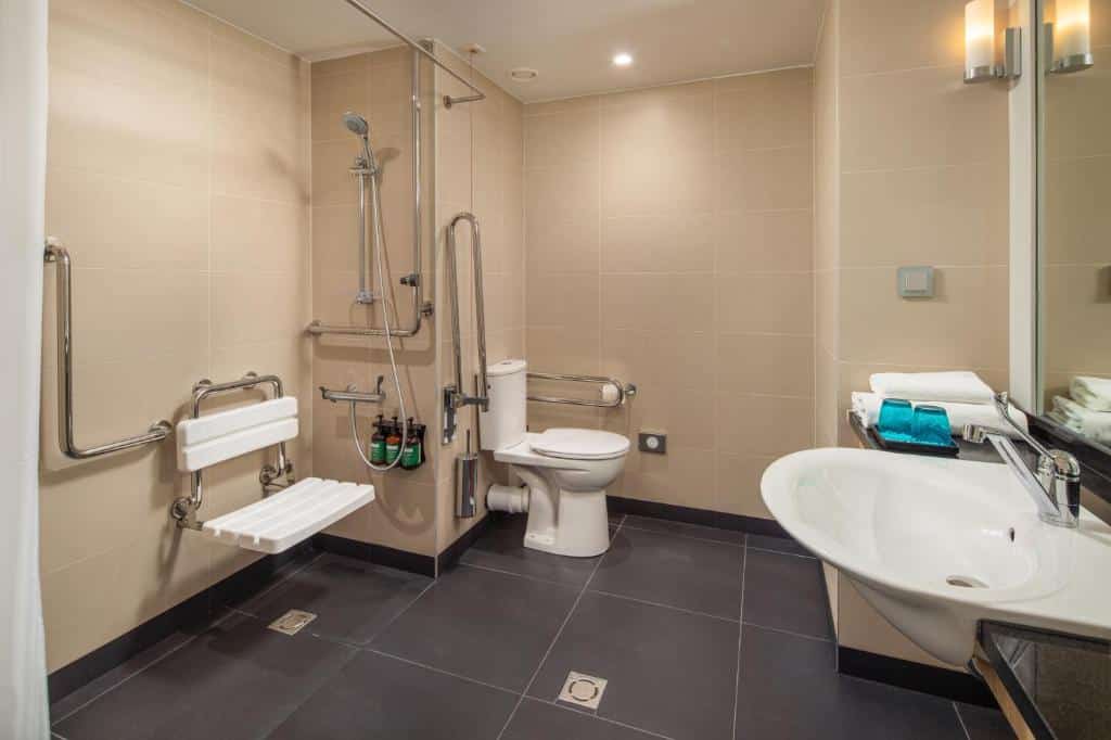 Banheiro acessível do Crowne Plaza Budapest by IHG com barras de apoio, assento de chuveiro montado na parede, vaso sanitário, pia e toalhas em um balcão com iluminação clara.