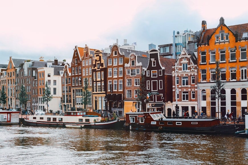 Fileira de edifícios coloridos e estreitos e casas flutuantes ao longo de um canal sob um céu nublado em um cenário de Amsterdam.