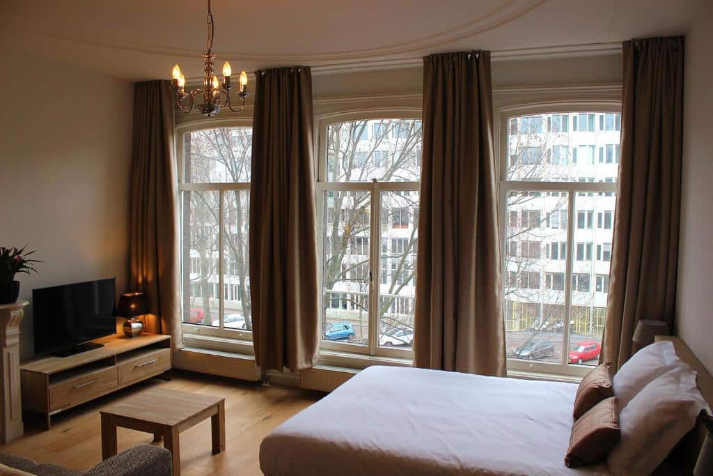 Foto do Amsterdam Jewel Canal Apartments que ilustra onde ficar em Amsterdam. Um quarto com uma cama grande, TV, móveis de madeira e três janelas altas com cortinas marrons com vista para uma rua.