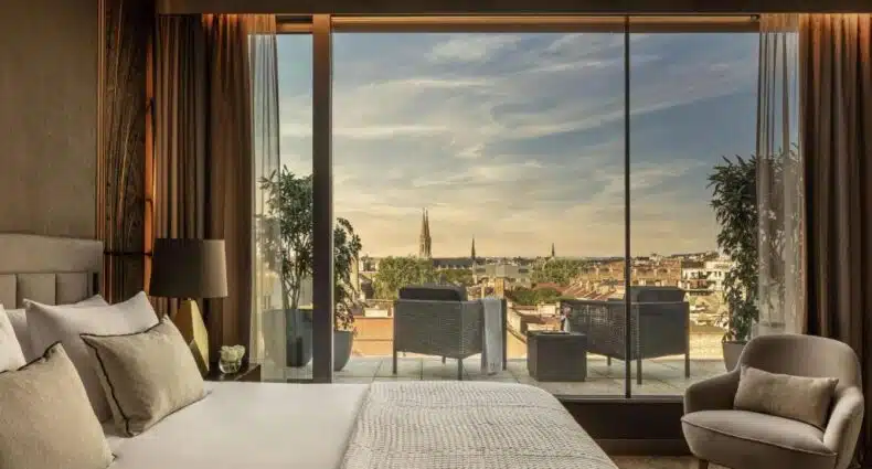 Quarto do Anantara New York Palace Budapest com uma cama grande, poltrona e varanda com vista para a cidade através de janelas do chão ao teto. Imagem para ilustrar post sobre onde ficar em Budapeste.