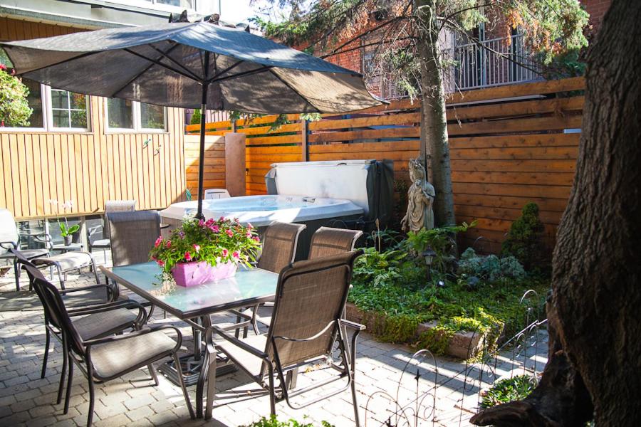 Pátio do Maison Des Jardins B & B com mesa de vidro, cadeiras, guarda-sol, vasos de flores, banheira de hidromassagem e área de jardim perto de uma cerca de madeira. Imagem para ilustrar post sobre onde ficar em Montreal.