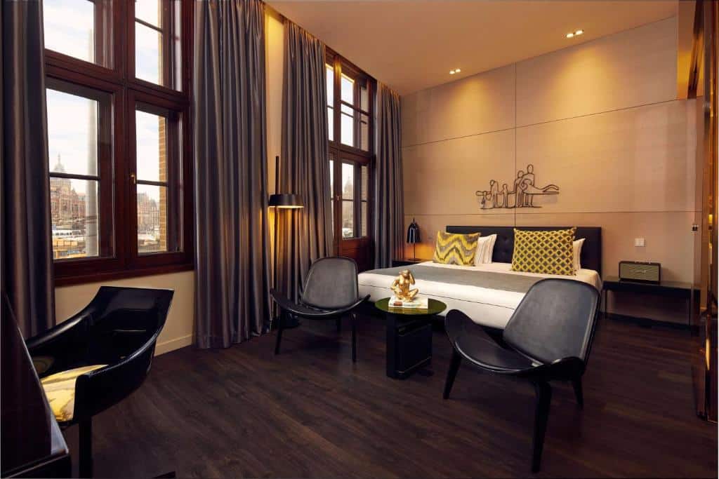 Foto do art’otel amsterdam que ilustra onde ficar em Amsterdam. Quarto de hotel moderno com uma cama grande, cadeiras, piso de madeira escura, janelas altas e cortinas cinza.
