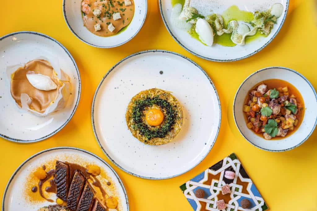 Foto do art’otel amsterdam. Seis pratos com variedades de pratos gourmet dispostos em uma mesa amarela brilhante, vistos de cima.