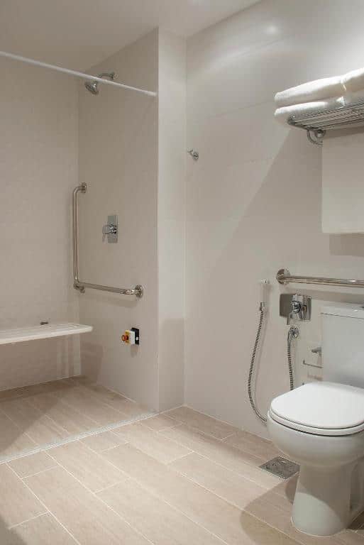 Banheiro acessível d Bella Gramado com assento de chuveiro montado na parede, barras de apoio, chuveiro de mão, vaso sanitário e toalheiro.