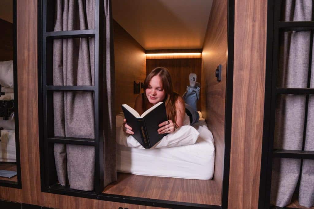 Foto do Cabins Hotel Amsterdam que ilustra onde ficar em Amsterdam. Uma pessoa deitada em uma cama em um quarto de cápsula de madeira, lendo um livro com cortinas cinza parcialmente fechadas.