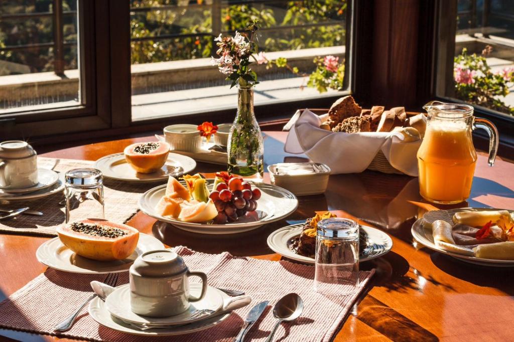Mesa de café da manhã do Hotel São Gotardo com frutas, pães, suco, xícaras de café, pratos, copos e um vaso de flores ao lado de uma janela ensolarada.