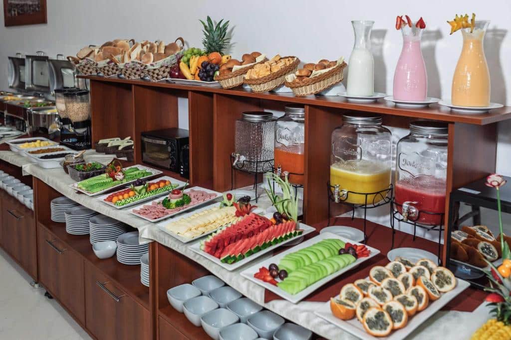 Uma mesa de bufê com frutas variadas, legumes, pães, bolos, cereais, distribuidores de suco e garrafas de leite no Quechua Hotel Cusco.