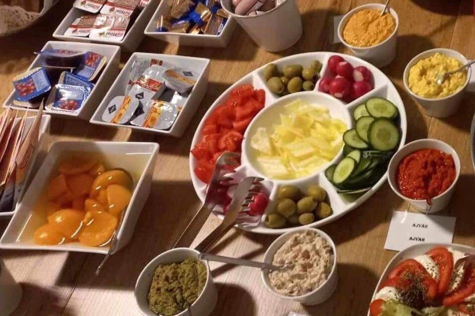 Uma mesa do Váralja Home com vários itens de café da manhã, incluindo vegetais fatiados, azeitonas, pastas, queijos e pêssegos em conserva.