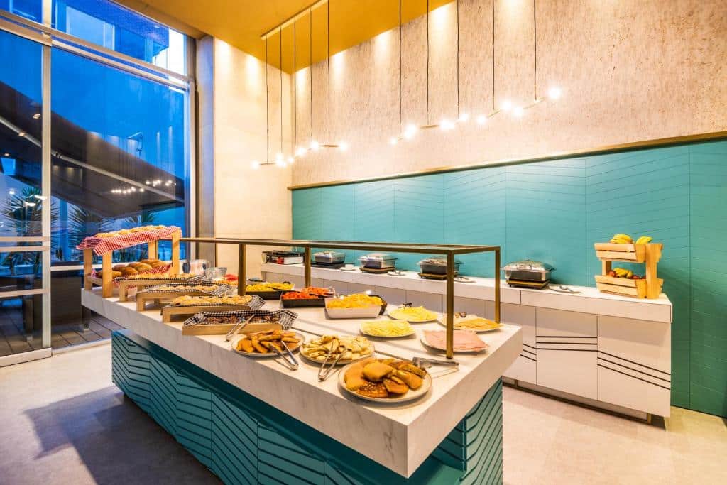 Uma mesa de bufê do ibis Bogota Museo com vários pães, frutas e pratos quentes em uma área de jantar moderna e bem iluminada, com decoração em azul-petróleo e branco. Imagem para ilustrar post sobre onde ficar em Bogotá.