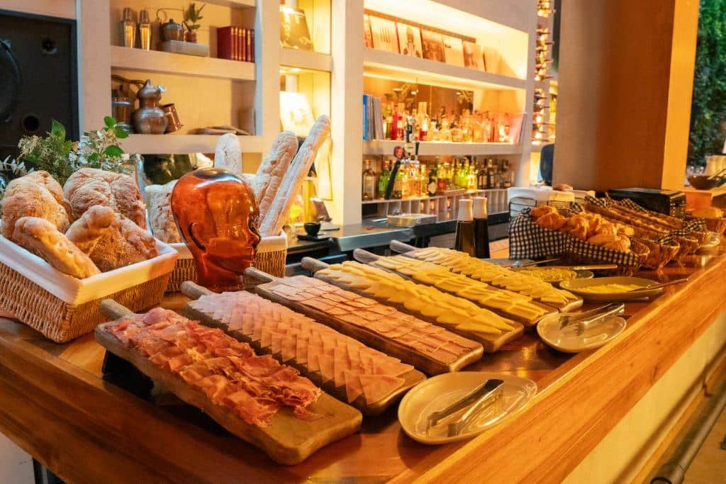 Um balcão de madeira do The Click Clack Hotel exibe pães variados, carnes fatiadas e queijos com pratos e utensílios em uma área de bar com iluminação aconchegante.