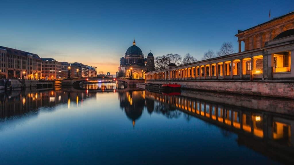 Uma paisagem urbana ao anoitecer com um edifício abobadado, um rio, pontes e edifícios iluminados refletindo na água.