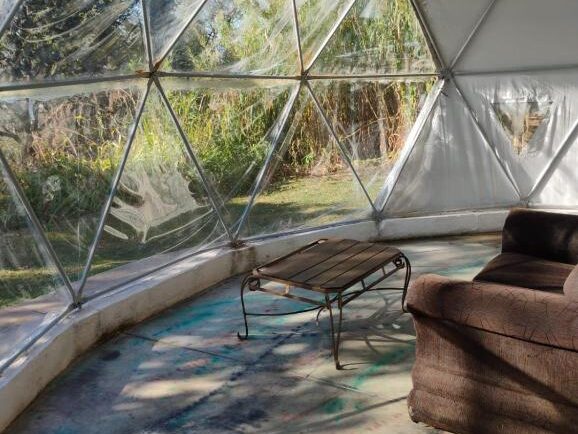 Uma poltrona marrom e uma mesa de centro de metal ficam dentro de uma cúpula da Pousada Spa Saúde Melhor, em Extrema, com janelas triangulares, com vista para a vegetação do lado de fora.