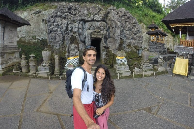 Um casal posa para uma selfie em frente à entrada de pedra do templo Goa Gajah em Bali, Indonésia. Representa onde ficar em Ubud