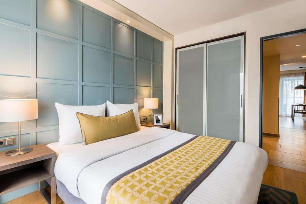 Foto do Citadines City Centre Frankfurt. Quarto moderno com uma cama grande, duas mesas de cabeceira com luminárias e portas de armário deslizantes que levam a um corredor.