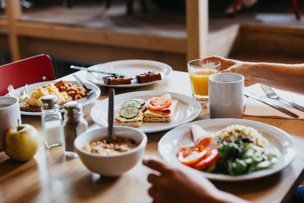 Foto do ClinkNOORD Hostel. Uma mesa posta com alimentos para o café da manhã, incluindo torradas, frutas, saladas, cereais, café e uma mão segurando um copo de suco de laranja.