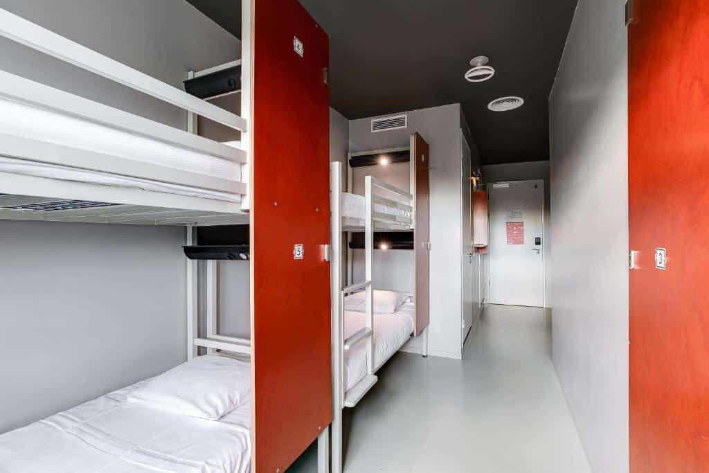 Foto do ClinkNOORD Hostel que ilustra onde ficar em Amsterdam. Quarto estreito com dois conjuntos de beliches, roupa de cama branca, armários vermelhos e uma porta no final do corredor.