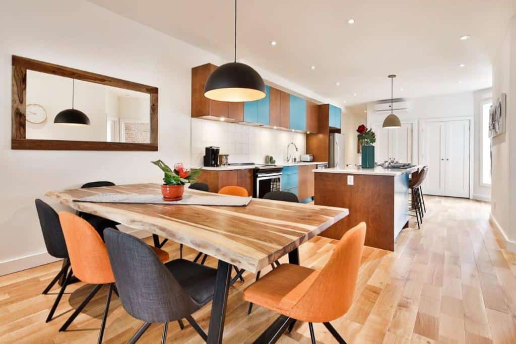 Cozinha aberta e área de jantar do Les Lofts du Mile End-Plateau Mont Royal com mesa de madeira, cadeiras de cores variadas, iluminação moderna e piso de madeira clara.