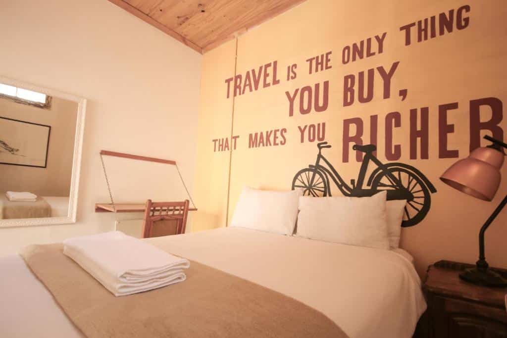 Uma cama de quarto no CURIOCITY Backpackers Johannesburg bem arrumada com roupas de cama brancas, um abajur, um gráfico de bicicleta e uma citação de viagem pintada na parede atrás.