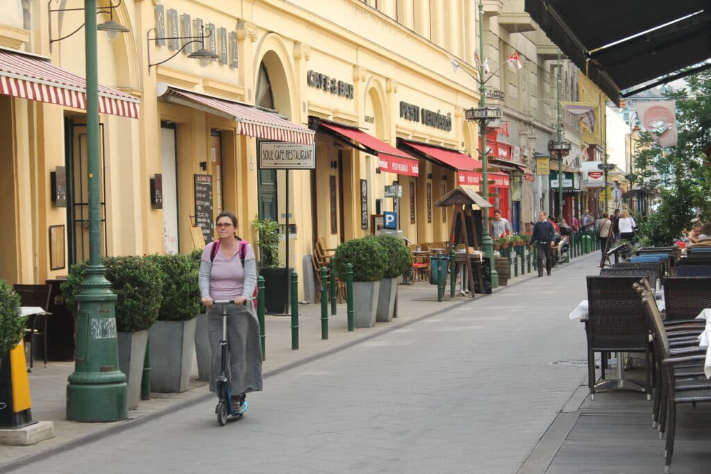 Uma mulher dirige uma scooter elétrica em uma rua de pedestres em Ferencváros repleta de cafés e restaurantes com mesas ao ar livre.