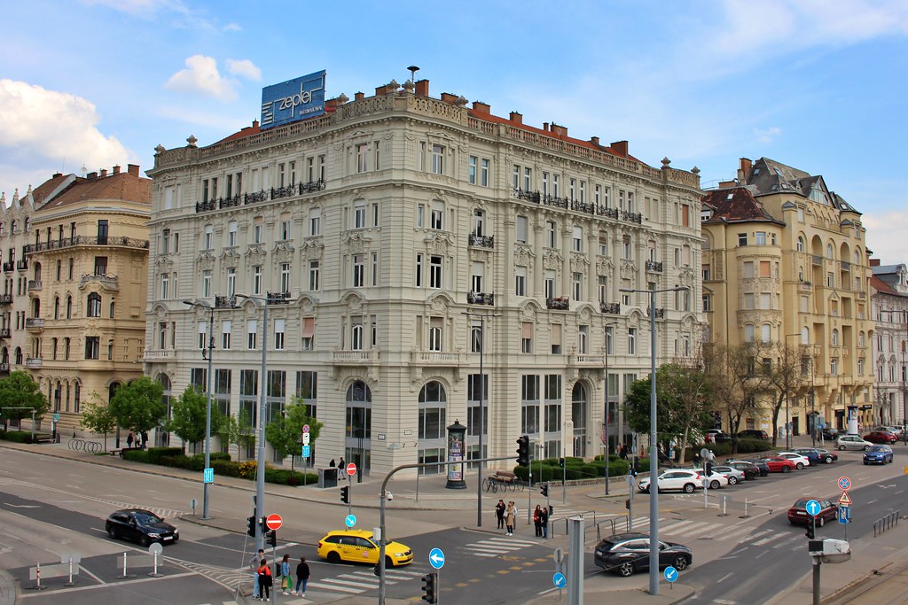 Um prédio grande em Belváros, ornamentado e de cor clara fica em um cruzamento movimentado da cidade com carros, pedestres e placas de rua.
