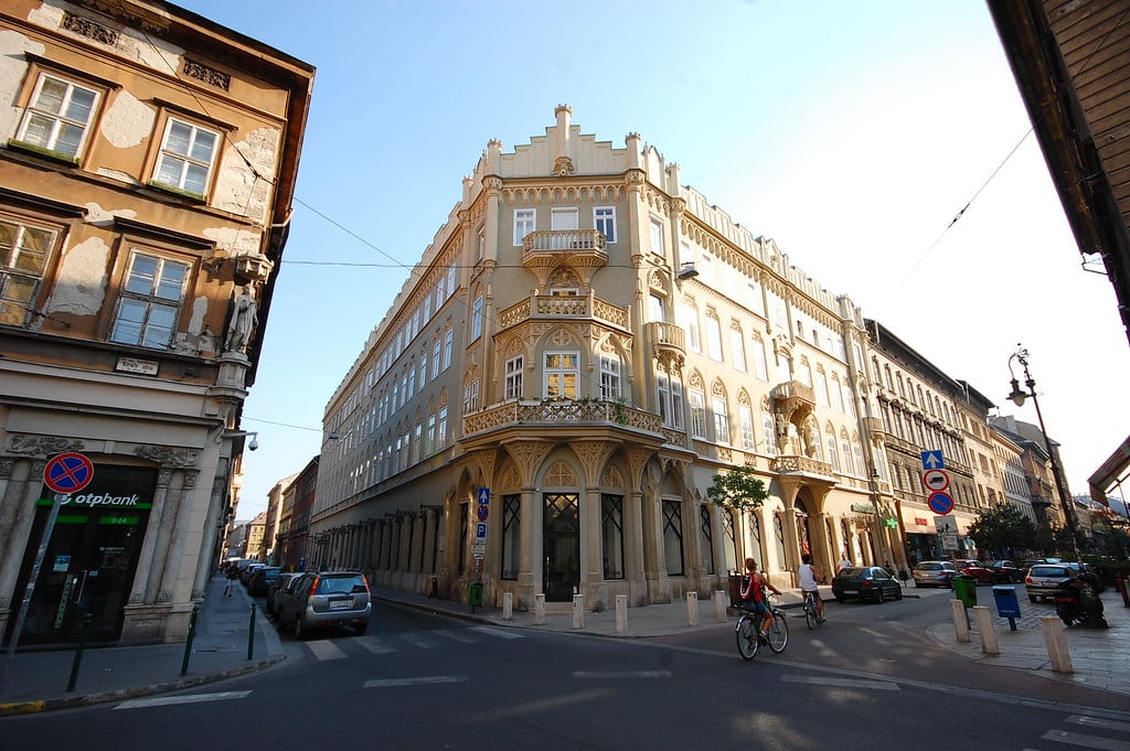 Vista de esquina de um edifício bege ornamentado em um cruzamento da cidade com carros estacionados e uma pessoa andando de bicicleta, em Terézváros. Imagem para ilustrar post sobre onde ficar em Budapeste.
