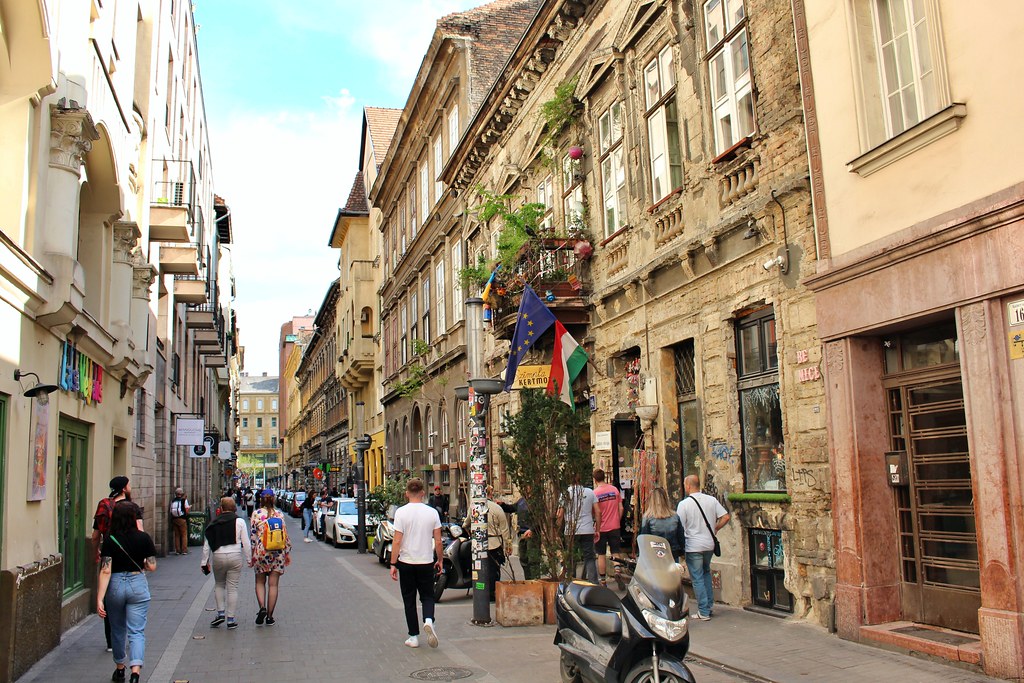 As pessoas caminham por uma rua estreita repleta de prédios antigos, lojas e veículos estacionados sob um céu parcialmente nublado, em Erzsébetváros.