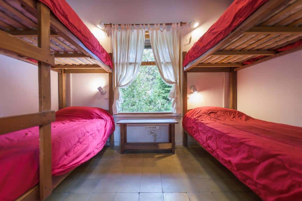 Um quarto com dois beliches de madeira, cada um com roupa de cama vermelha, e uma janela com cortinas brancas entre eles.