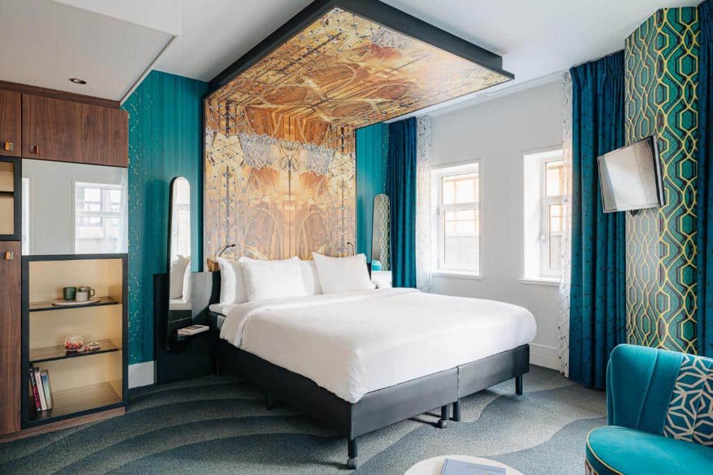 Foto do Eden Hotel Amsterdam que ilustra onde ficar em Amsterdam. Quarto de hotel com uma cama grande, decoração em azul-petróleo e dourado, arte na parede acima da cama, duas janelas e uma TV na parede.