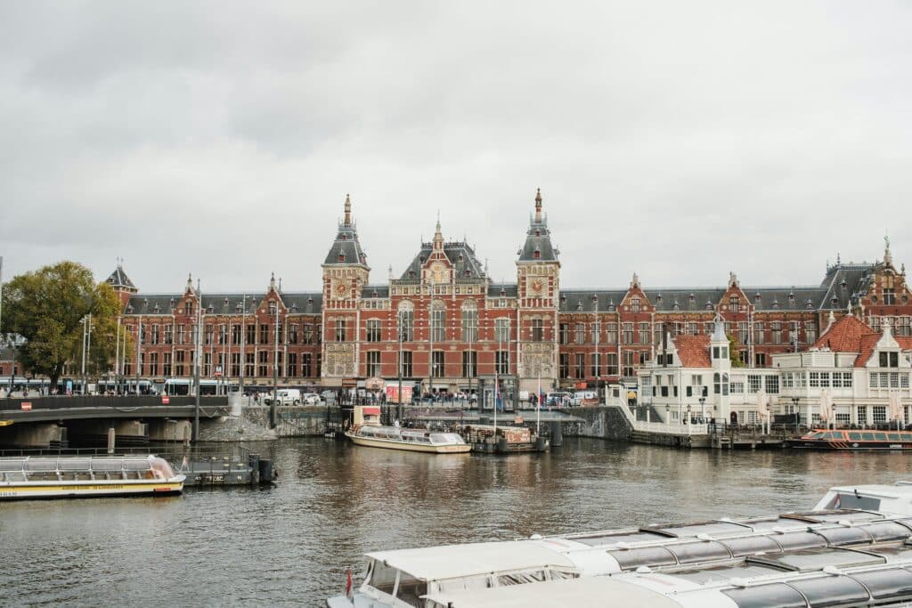 Grande edifício de tijolos vermelhos com torres em um canal, barcos atracados ao longo da orla e céu nublado.