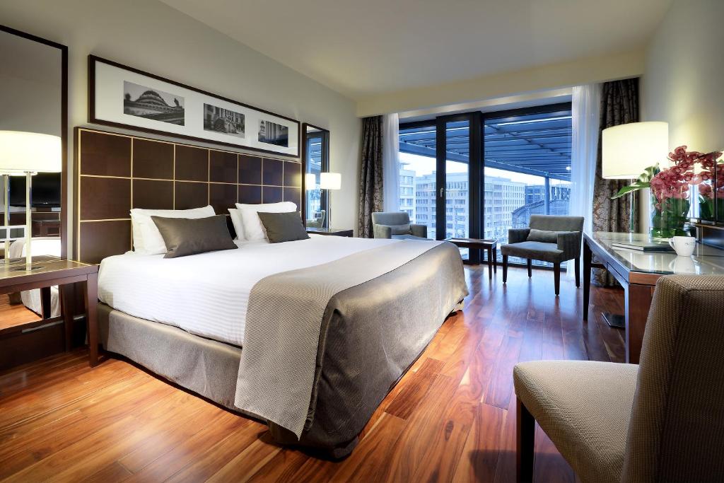Quarto de hotel moderno no Eurostars Berlin com uma cama grande, poltronas, escrivaninha, luminárias, piso de madeira e vista da cidade através de grandes janelas. Está ilustrando onde ficar em Berlim.