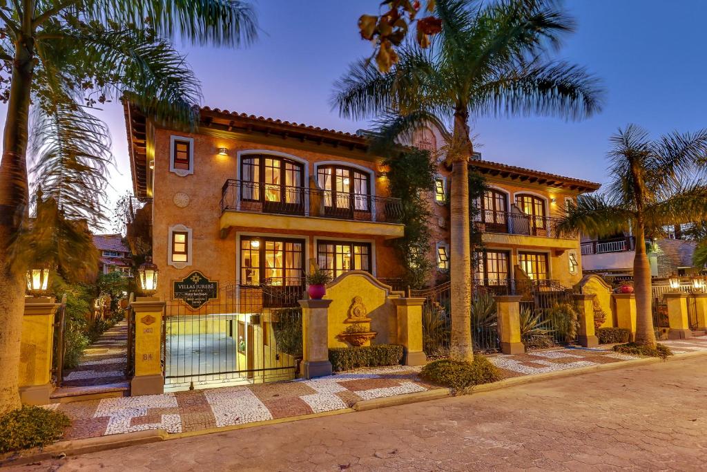 Fachada do Villas Jurerê Hotel Boutique, uma villa de três andares em estilo mediterrâneo com varandas, iluminação externa quente, palmeiras e uma entrada fechada ao anoitecer.