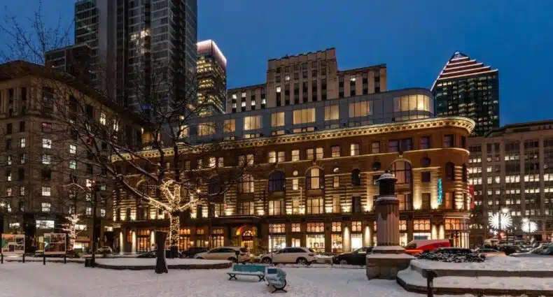 Um parque urbano coberto de neve ao anoitecer com bancos, árvores sem folhas e edifícios históricos e modernos iluminados ao fundo. Imagem da fachada do Hôtel Birks Montréal para ilustrar post sobre onde ficar em Montreal.
