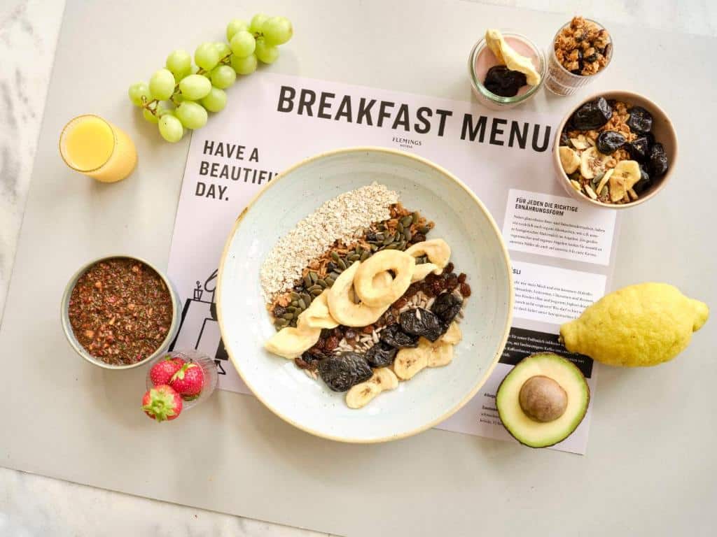 Foto do Flemings Hotel Frankfurt-Central. Uma tigela de aveia com frutas e nozes, rodeada de uvas, limão, abacate, morangos, suco e cardápio de café da manhã.