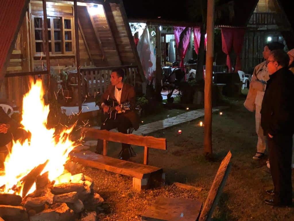 Um homem toca violão ao lado de uma fogueira à noite na Pousada e Camping Tio Wil, enquanto outras pessoas ficam por perto, com cabanas e velas acesas ao fundo.