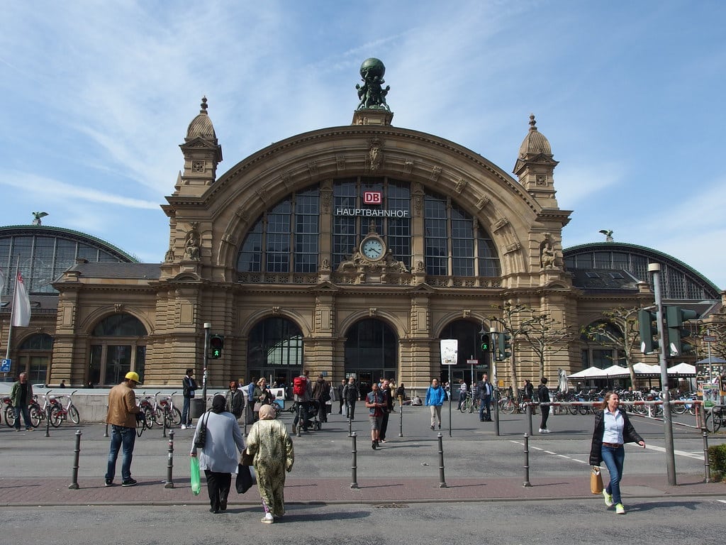 Pessoas caminham em frente à entrada principal da Frankfurt Hauptbahnhof, uma grande estação de trem histórica na Alemanha.