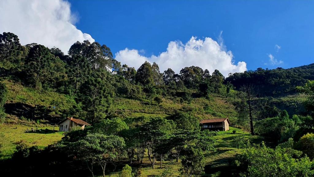 Duas pequenas casas em uma encosta gramada cercada por árvores sob um céu azul com nuvens dispersas, na Pousada Sítio Nova União. Representa hotéis e Pousadas em Itamonte.