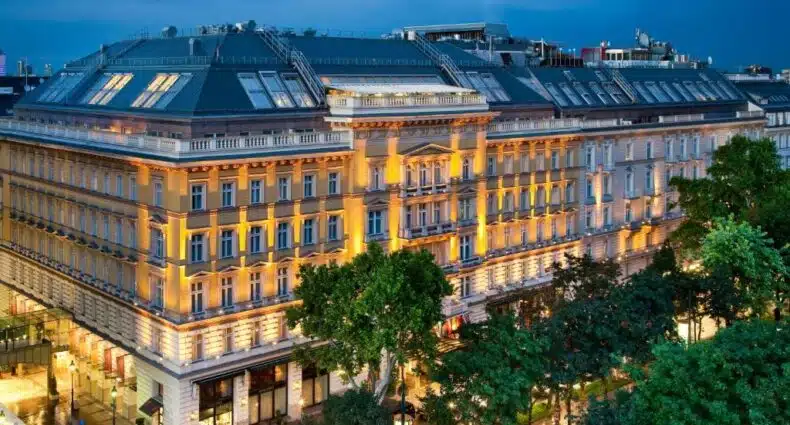 Grande edifício histórico com arquitetura ornamentada, iluminado ao anoitecer, com árvores e lojas ao longo da rua em frente, trata-se do Grand Hotel Wien, para representar onde ficar em Viena.