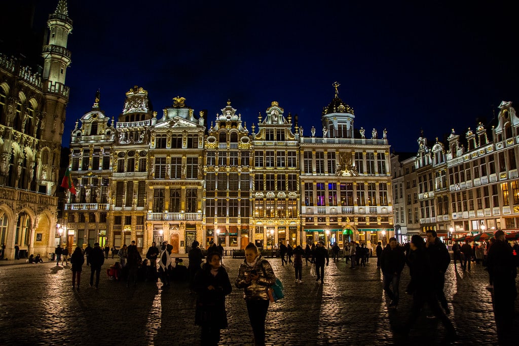 As pessoas caminham por uma Grand Place à noite, com edifícios ornamentados e iluminados ao fundo.