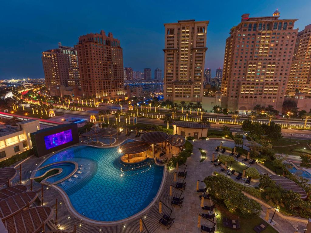 Vista da área da piscina do Hilton Doha The Pearl com espreguiçadeiras, cercada por prédios altos e luzes da cidade ao anoitecer.
