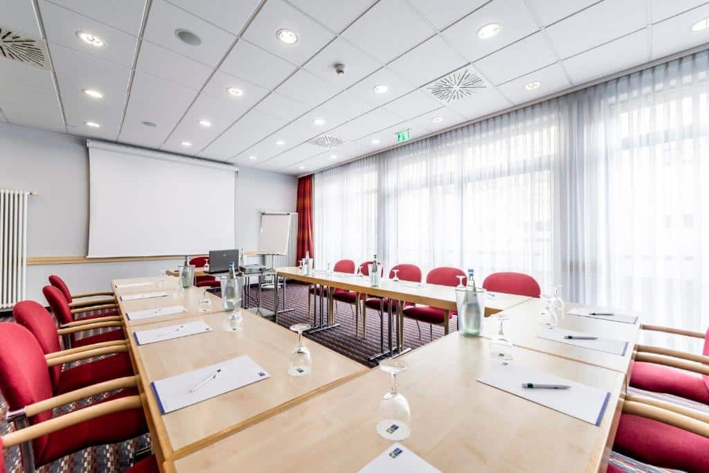Sala de conferência no Holiday Inn Express Berlin com cadeiras vermelhas, mesas em forma de U, blocos de anotações, copos, garrafas, tela de projetor e janelas grandes.