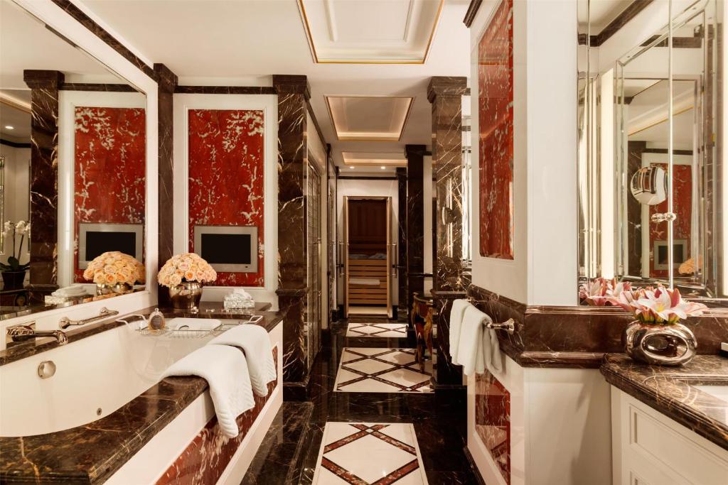 Hotel Adlon Kempinski Berlin. Banheiro luxuoso com piso de mármore, banheira grande, paredes com detalhes em vermelho e marrom, espelhos e flores frescas nos balcões.
