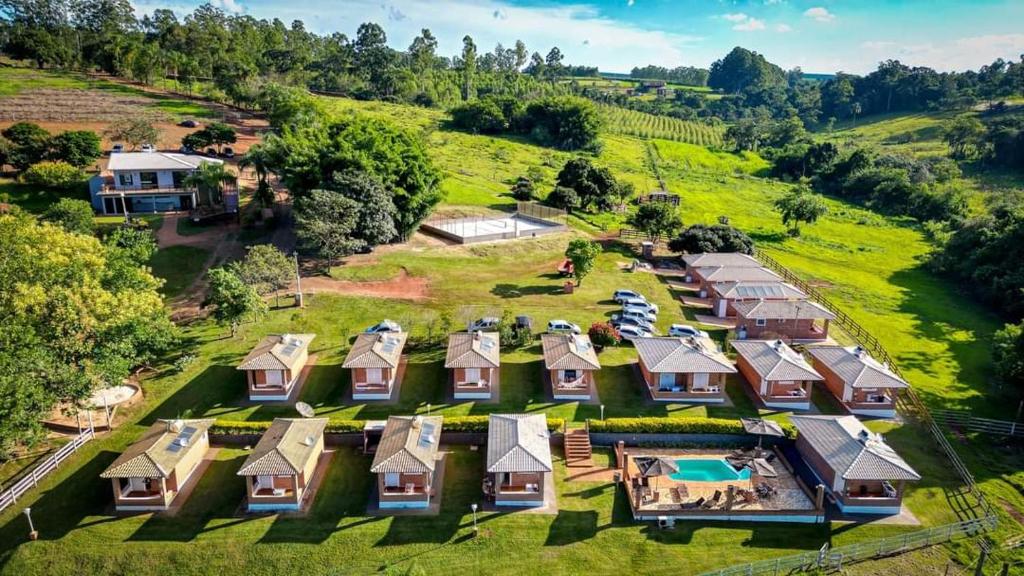 Vista aérea de pequenas cabanas, uma casa, piscina e carros em uma propriedade rural gramada, cercada por árvores e terrenos abertos.