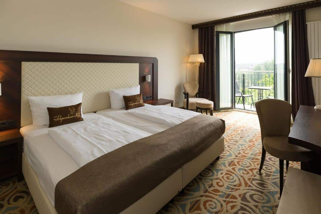 Foto do Hotel Schopenhauer Hof, representando onde ficar em Frankfurt. Quarto de hotel com uma cama de casal, cadeiras, escrivaninha e porta da varanda aberta que permite a entrada de luz natural.