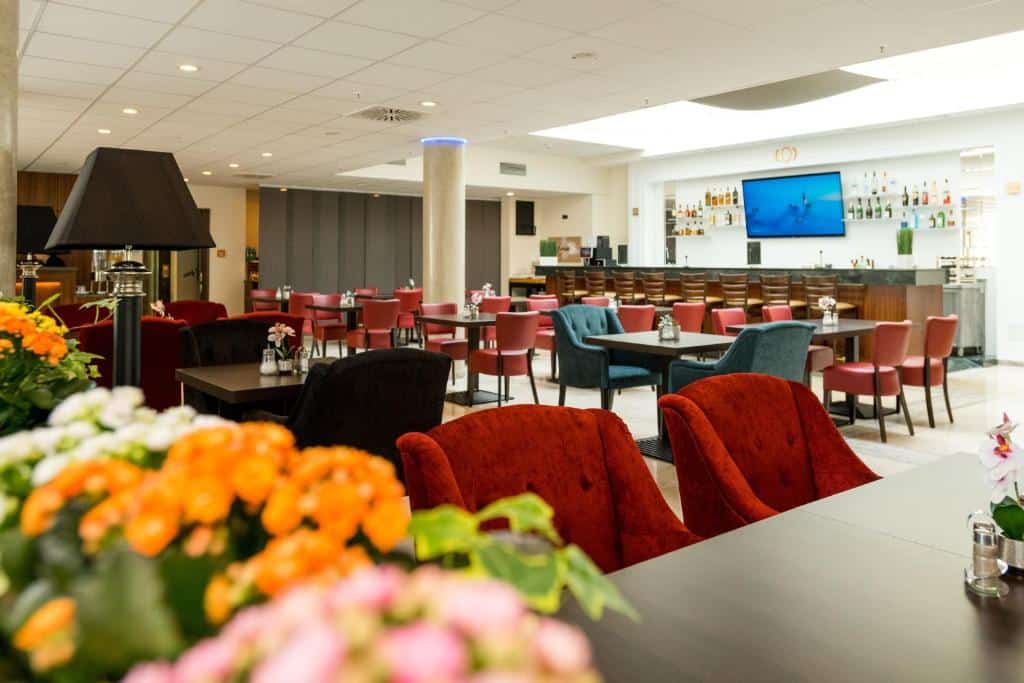 Foto do Hotel Schopenhauer Hof. Lounge moderno de hotel com cadeiras vermelhas e azuis, mesas, uma área de bar e uma televisão na parede; flores em primeiro plano.