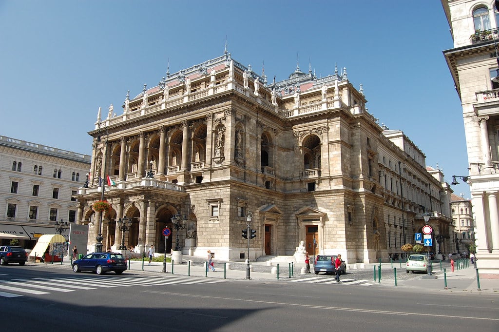 Um grande edifício de pedra ornamentado de Hungarian State Opera House com colunas e varandas fica em uma esquina da cidade sob um céu azul claro.